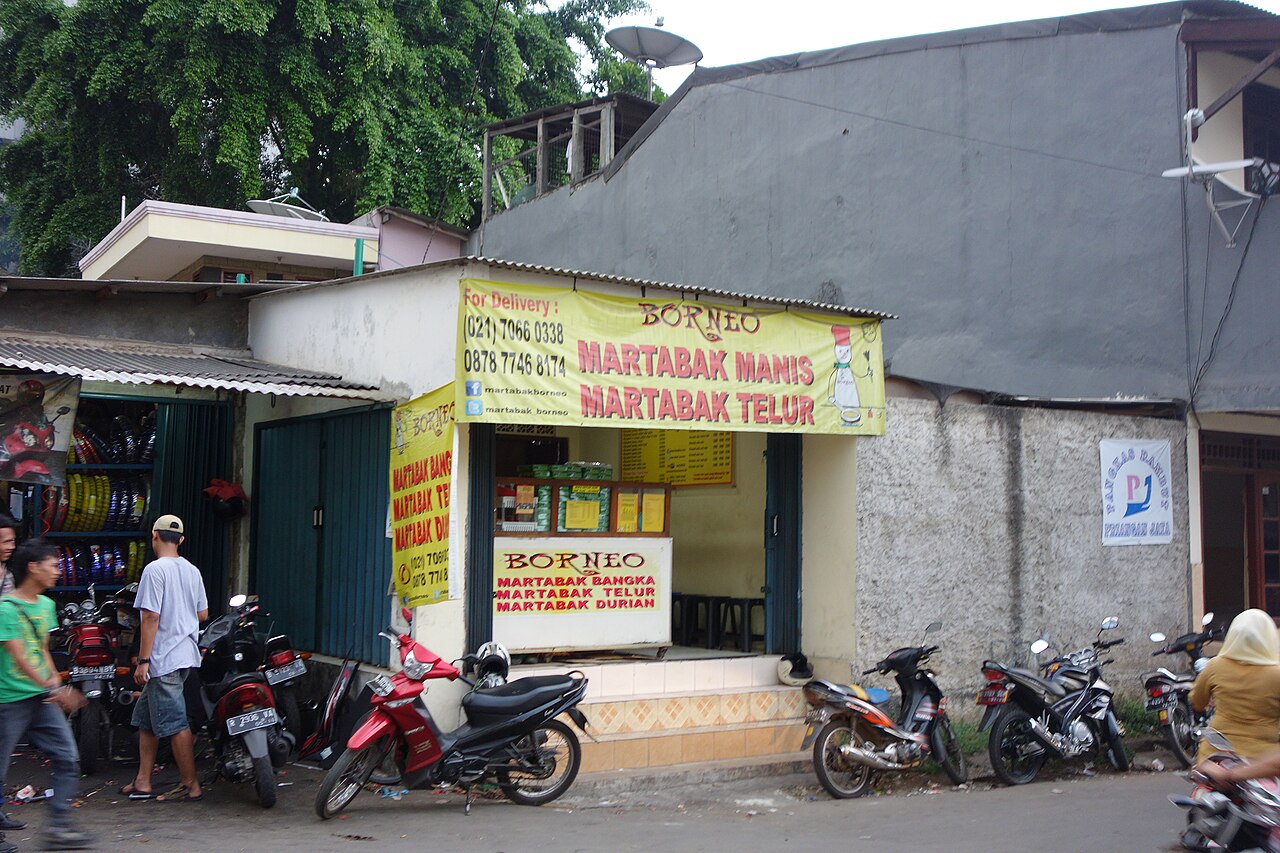 Warung martabak dengan spanduk kuning berisi nomor delivery dan akun media sosial, motor parkir di depannya